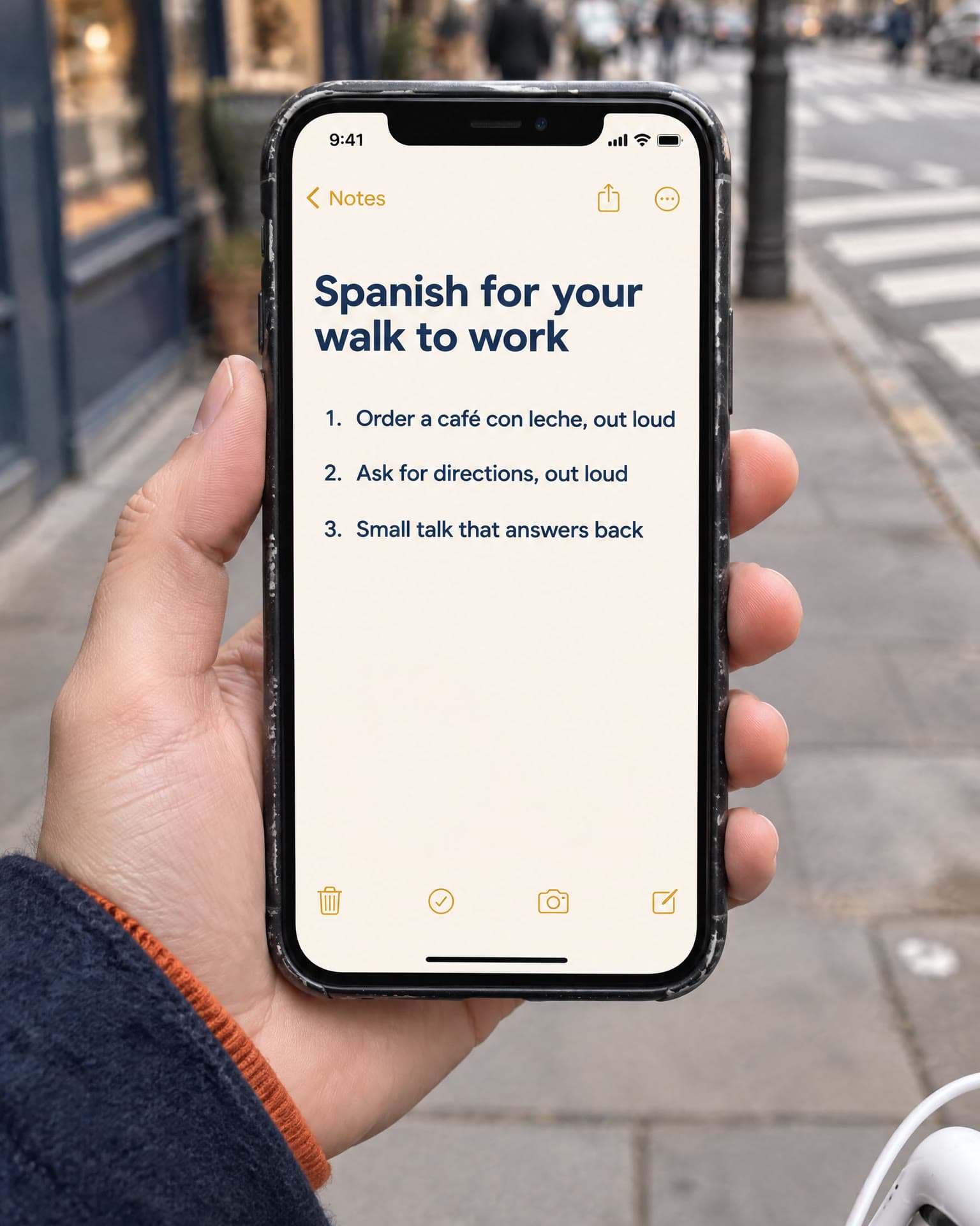 A hand holds a phone open on a notes page, on a city pavement. It reads: Spanish for your walk to work. Order a café con leche, out loud. Ask for directions, out loud. Small talk that answers back.