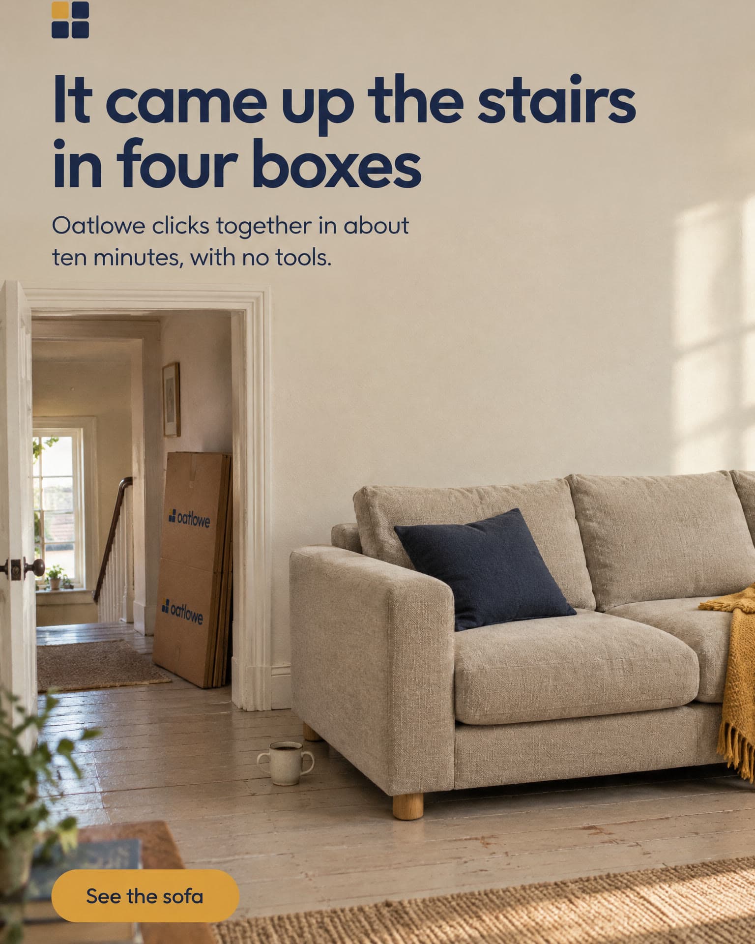 An oat sofa with a navy cushion in a sunny flat, flattened Oatlowe boxes leaning in the hallway behind it. It reads: It came up the stairs in four boxes. Oatlowe clicks together in about ten minutes, with no tools. See the sofa.